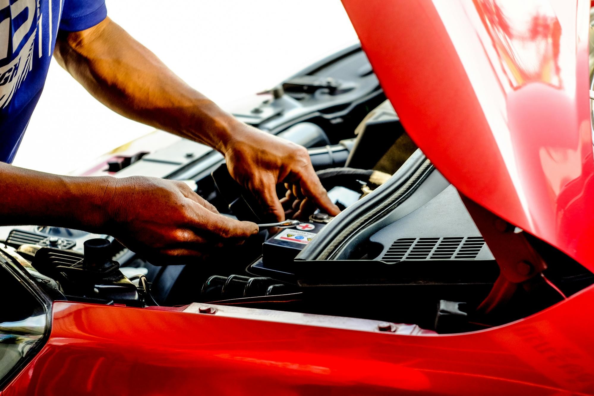 A person working on a car battery under the hood of a red car