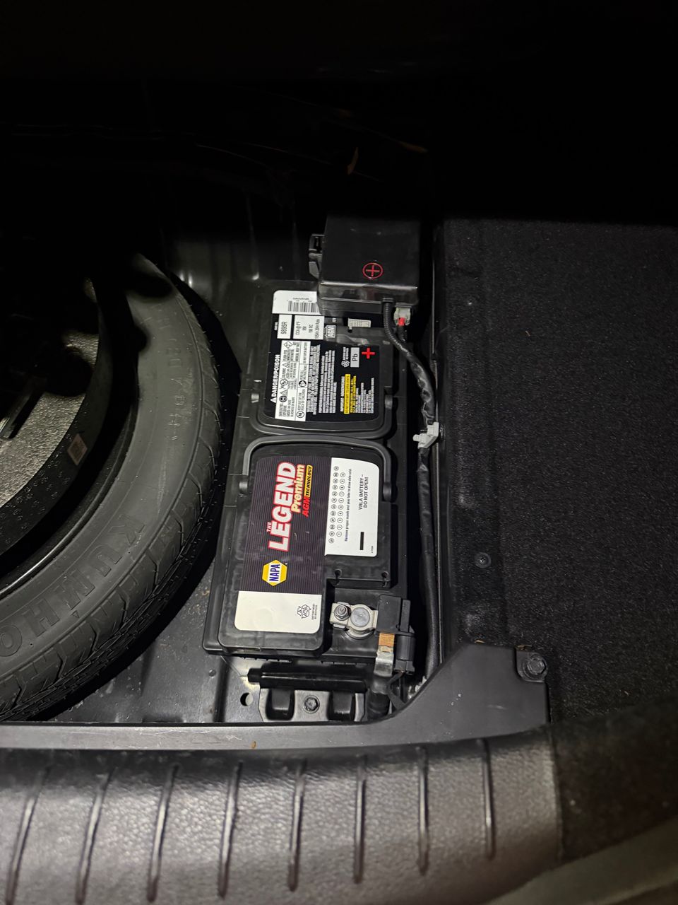 Old AGM car battery in a sedan's trunk compartment next to the spare tire, with a red plus mark on the terminal cover