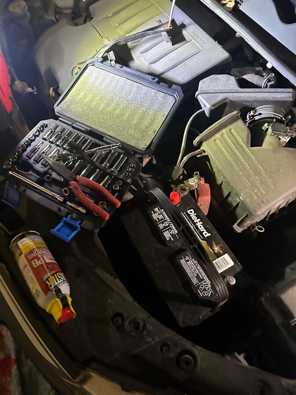 DieHard battery and an open socket set in a car's engine bay, lit by a work light after dark