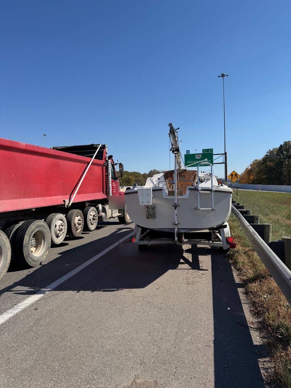 Sailboat on a trailer stopped on a narrow I-270 shoulder by the guardrail while a red dump truck passes close, with the exit 27 Cleveland Ave sign ahead
