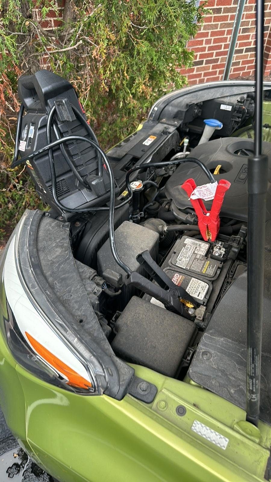 Portable booster pack resting on the open hood of a green hatchback, with red and black clamps on the battery