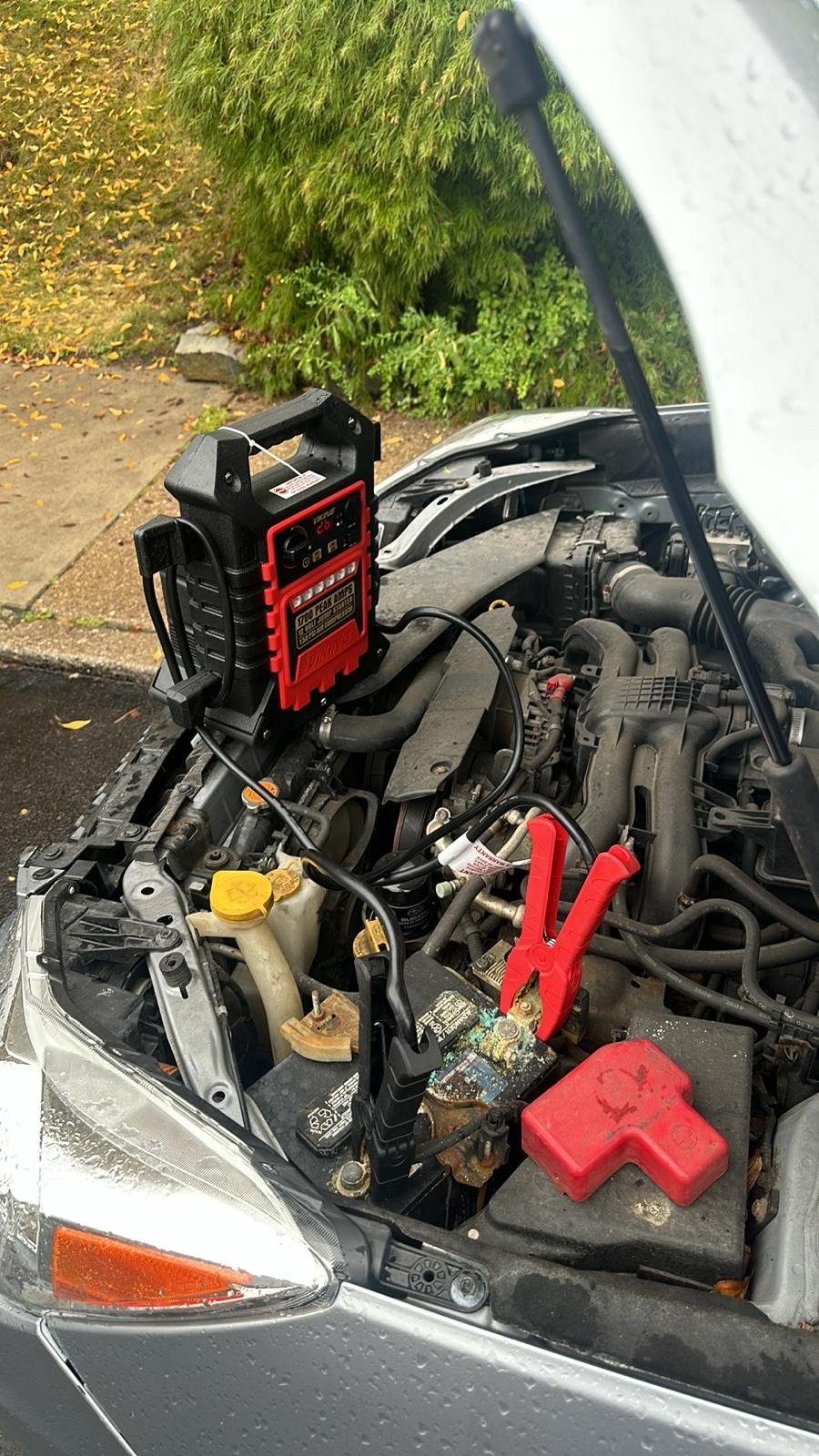 Booster pack on the engine of a silver car with its clamps on the battery, raindrops on the hood and headlight and wet pavement beside it