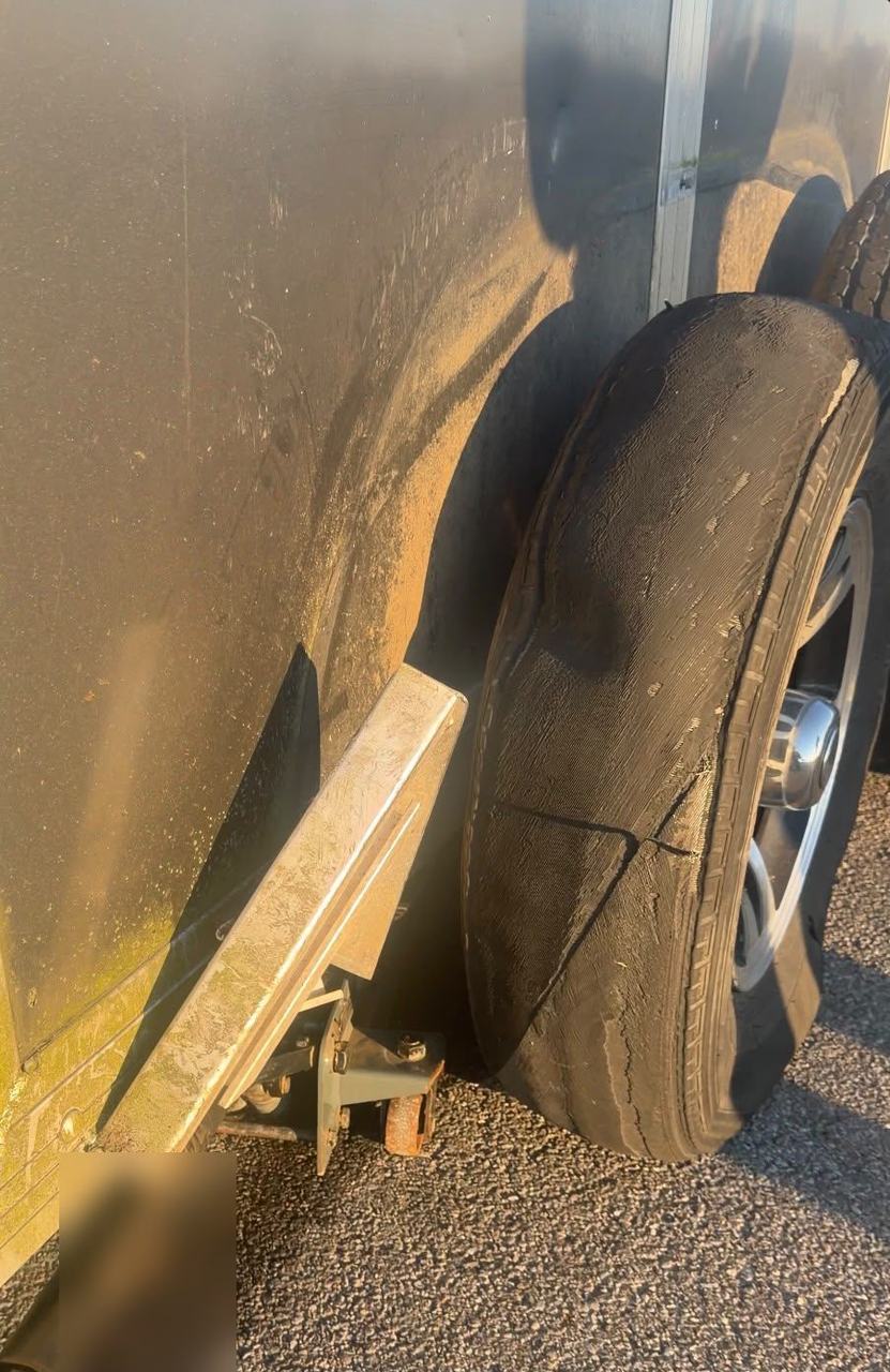 Cargo trailer tire with a shredded, worn-through tread after being driven flat, with a jack under the trailer frame