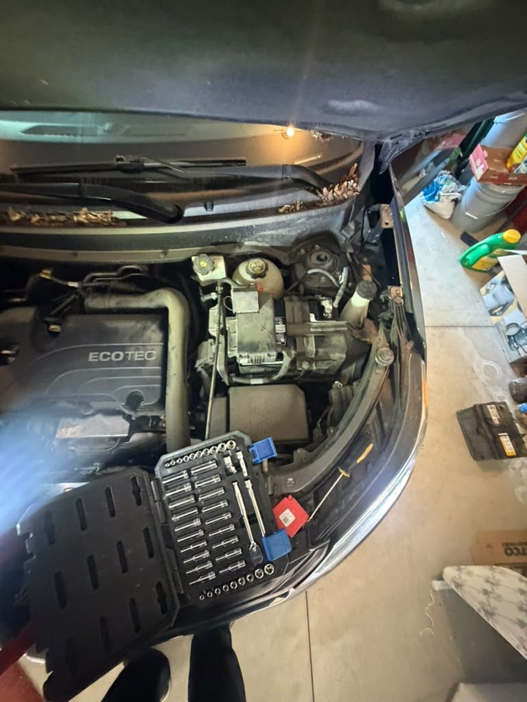 Open Chevrolet engine bay in a garage with a socket set on the bumper and the old battery on the floor