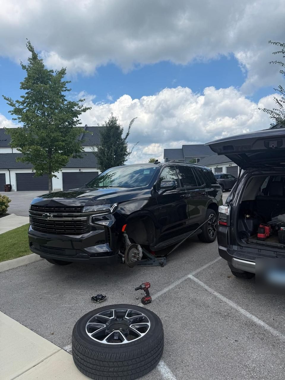 Chevrolet Tahoe on a floor jack in a parking lot with one wheel off and the tire lying tread side up, grey service truck alongside