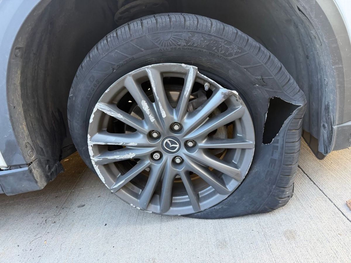 Gray alloy wheel on a white SUV with a piece of the rim broken out, curb scrapes on the spokes and a torn flat tire