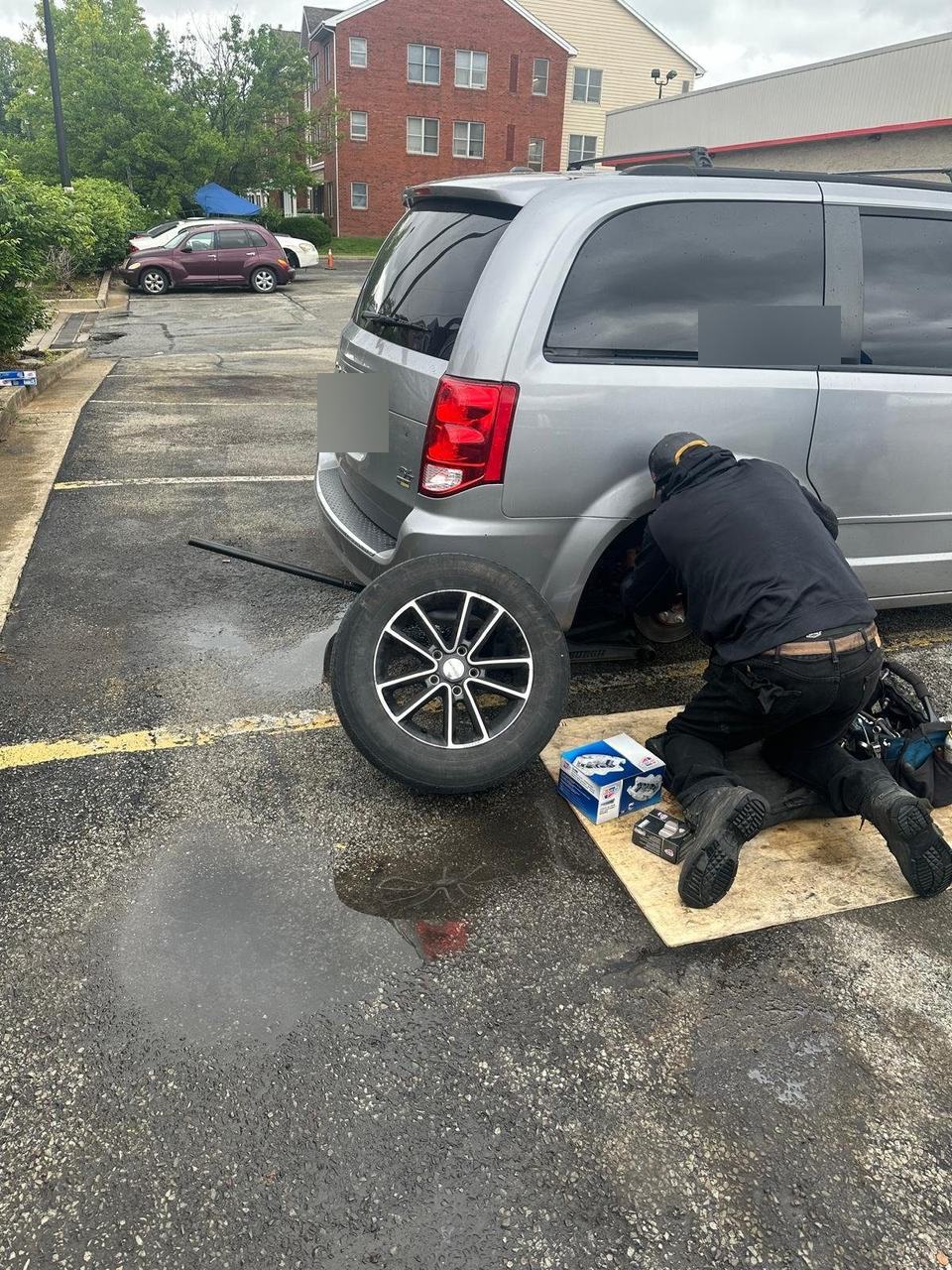 Technician kneeling at the rear hub of a silver Dodge Grand Caravan, the wheel off and leaning against the van