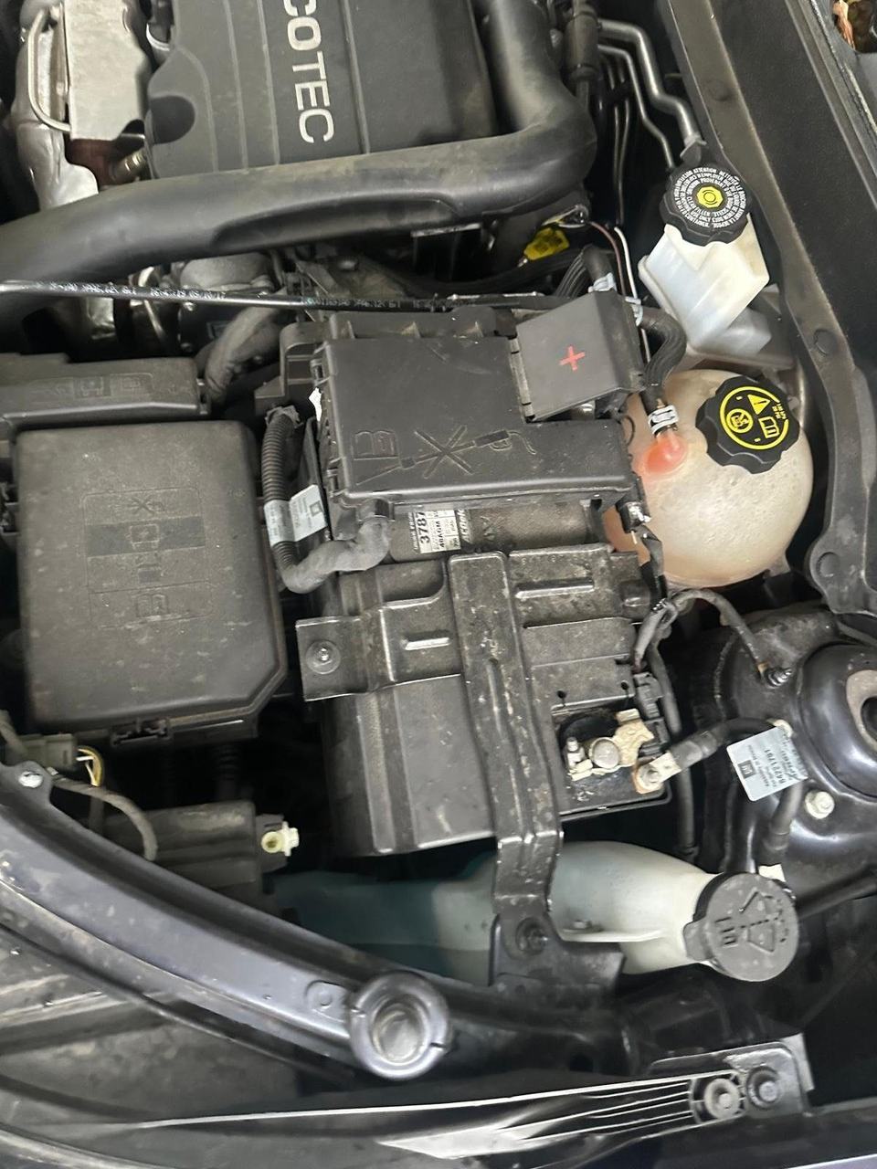 Battery under a plastic cover and hold-down bracket next to the coolant tank in an engine bay with an Ecotec engine