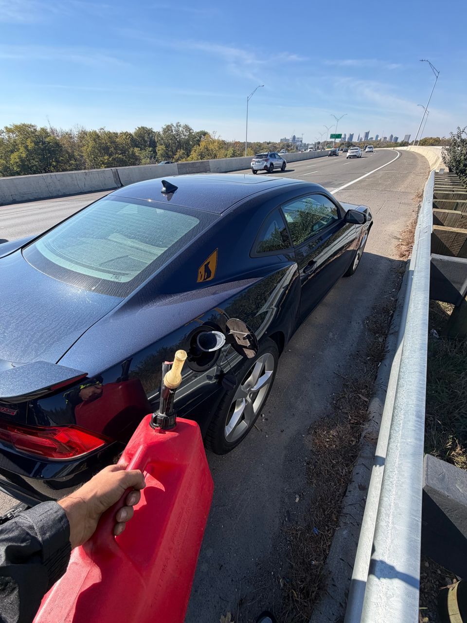 Red gas can held by the open fuel door of a dark blue Camaro on a narrow I-70 shoulder, with the downtown Columbus skyline ahead