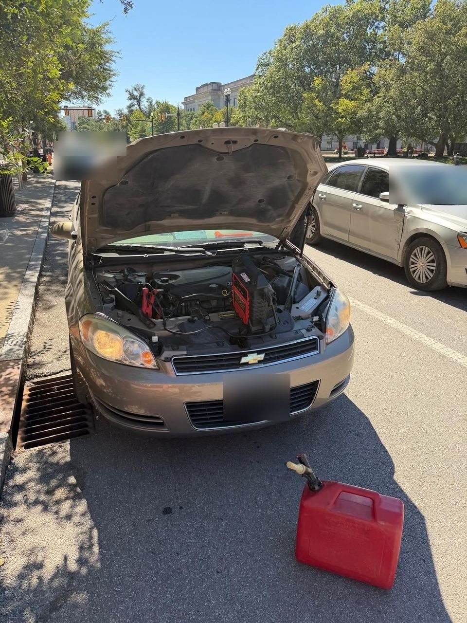 Red gas can on the pavement in front of a car with its hood up and a booster pack on the engine, parked at the curb