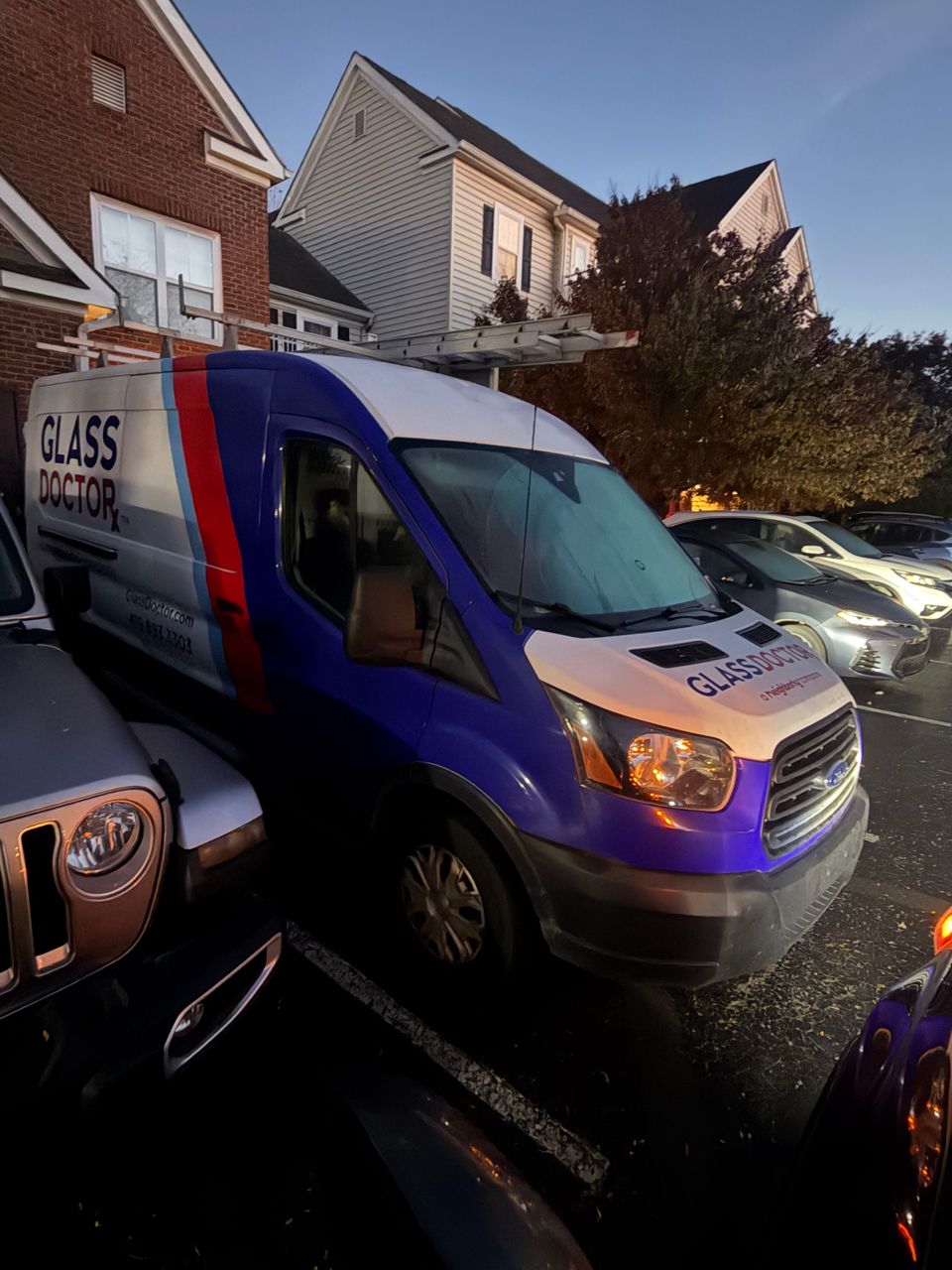 Glass Doctor Ford Transit work van with a ladder rack, parked in an apartment lot at dawn