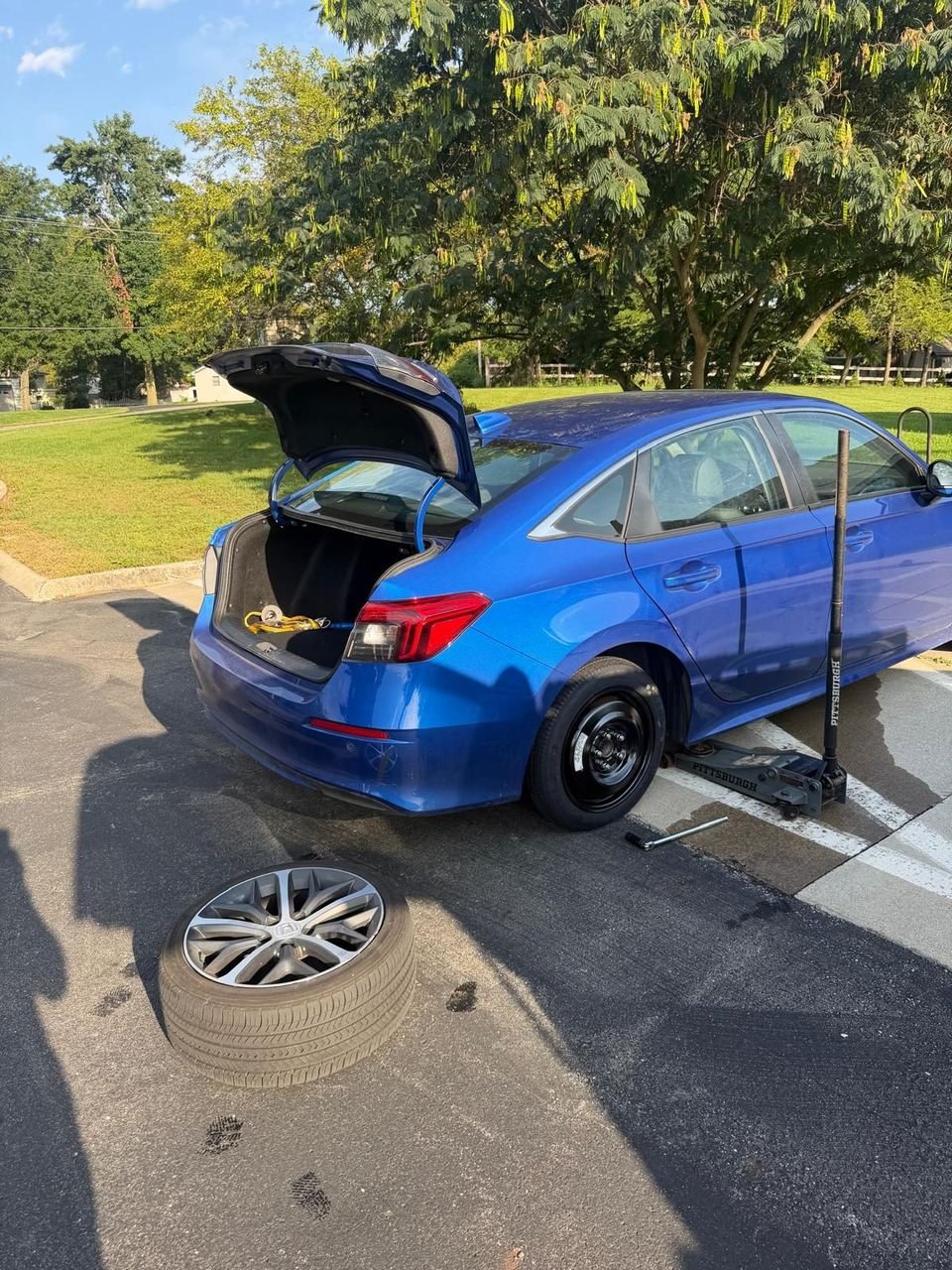 Blue Honda Civic on a floor jack with the spare fitted and the removed wheel lying on the pavement
