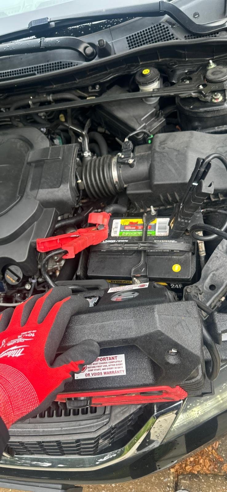 Red and black jump clamps on a battery in an engine bay, a gloved hand resting on the jump pack in front
