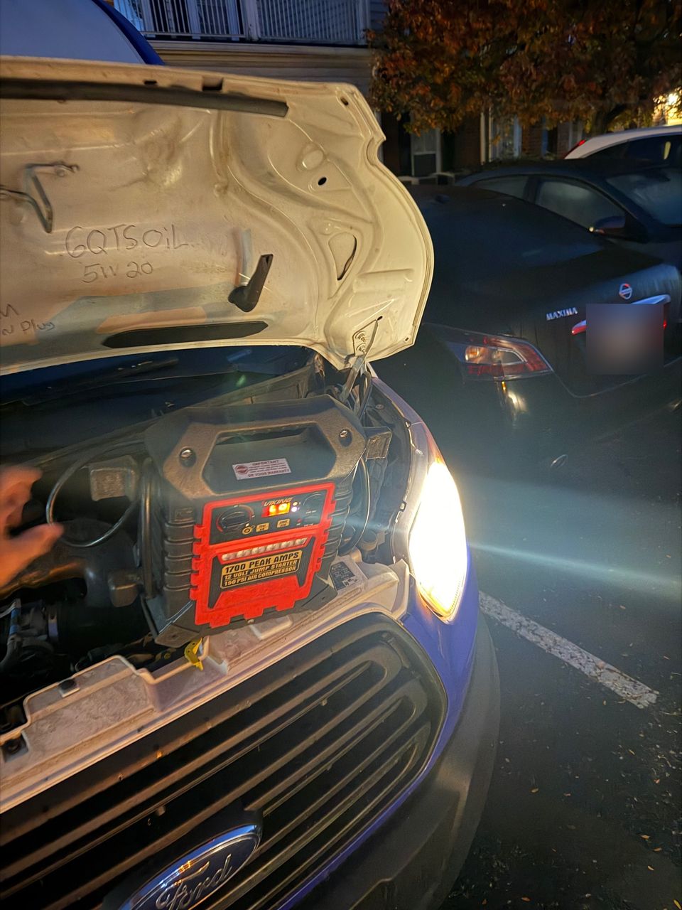 Red and black 1,700 peak amp jump pack sitting on the engine bay of a Ford Transit with the hood open, headlight on