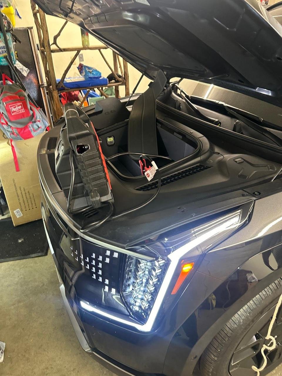 Jump pack resting on the front of a dark SUV in a home garage, its clamp cable running under the open hood