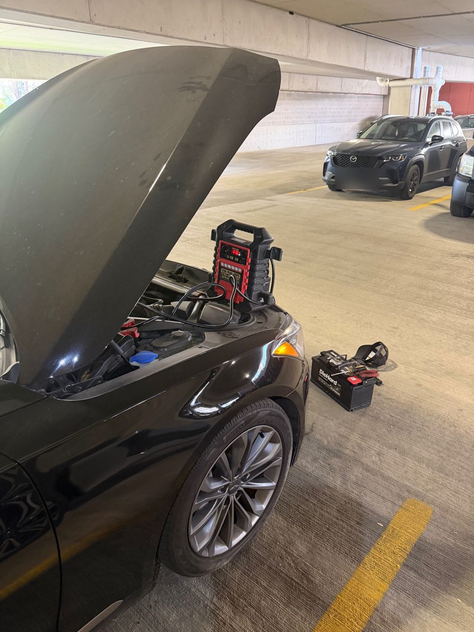 Black sedan in a parking garage with the hood up and a red portable jump pack clamped to the jump posts under the hood, a new DieHard battery on the floor