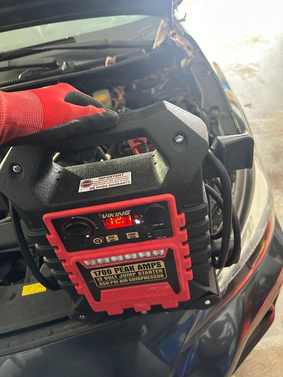 Technician in a red work glove holding a jump starter with a lit display in front of a car with the hood open