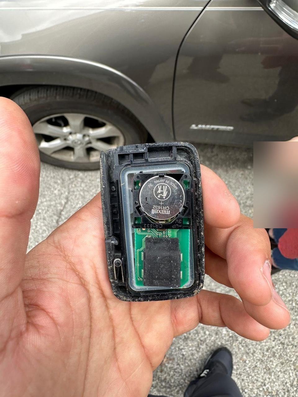 Hand holding an opened smart key fob showing a CR1632 coin cell, with a gray SUV behind