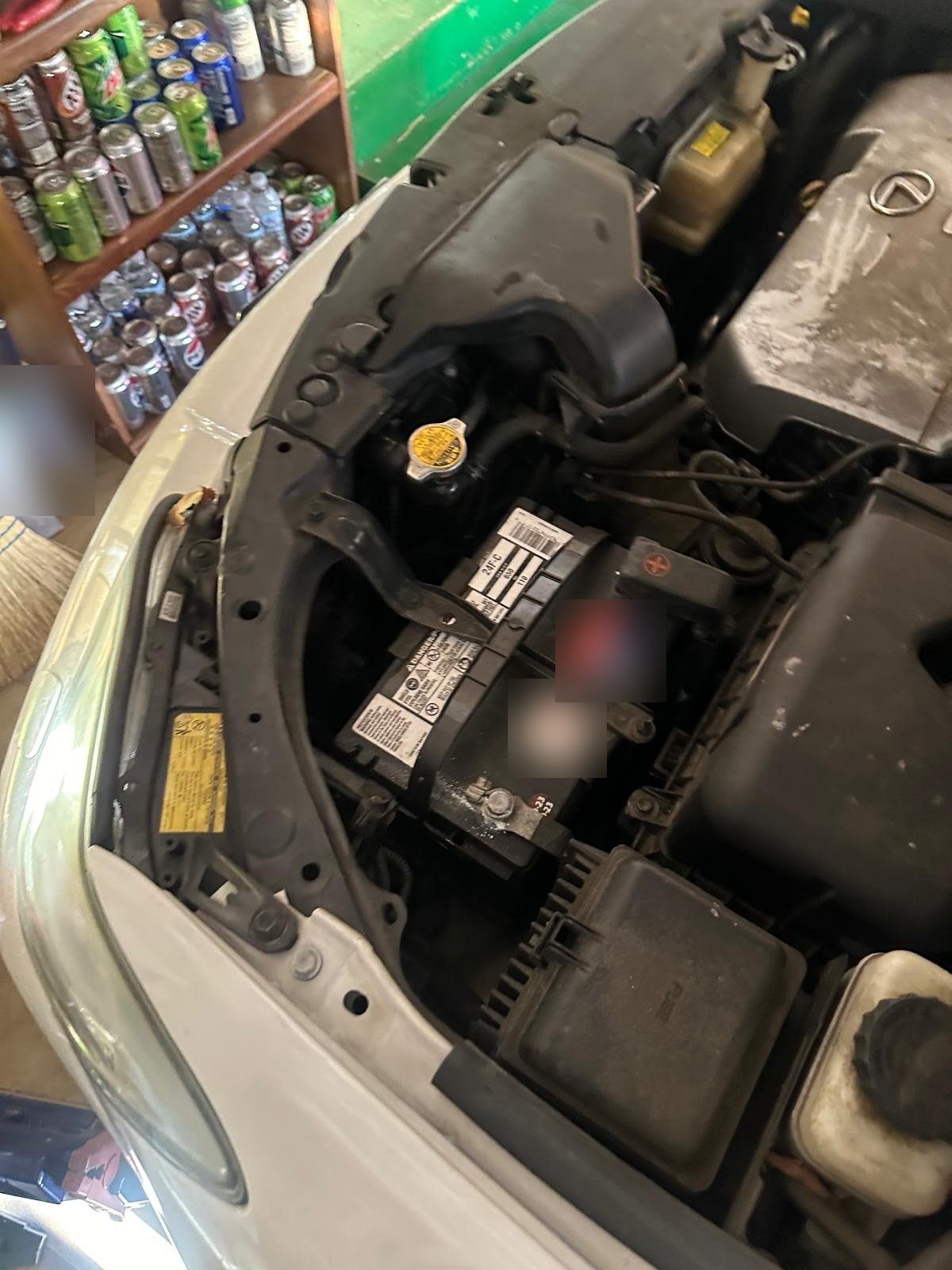Battery held in place by a metal hold-down bar in the engine bay of a white Lexus with the hood open