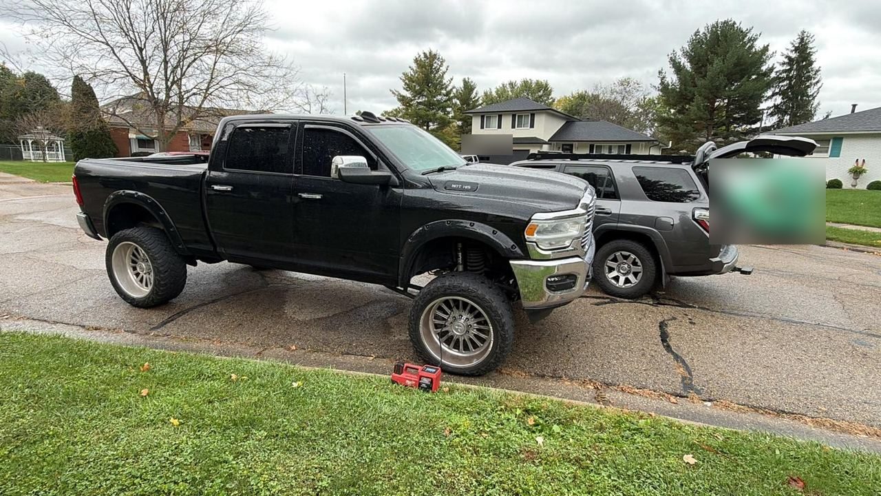 Red portable tire inflator on the curb beside the front tire of a black lifted pickup, with a grey Roadmate service truck parked behind