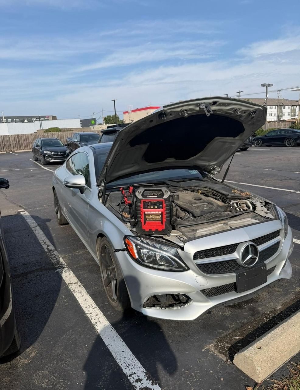 Red portable jump starter sitting in the engine bay of a silver Mercedes C-Class with the hood open in a parking lot