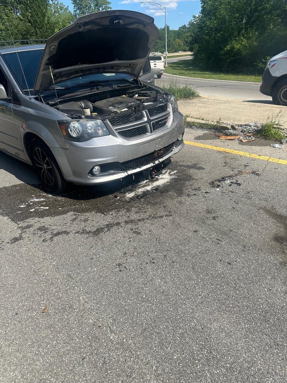 Silver minivan at the roadside with its hood open after a fire under the front bumper, burned cardboard and fluid on the pavement