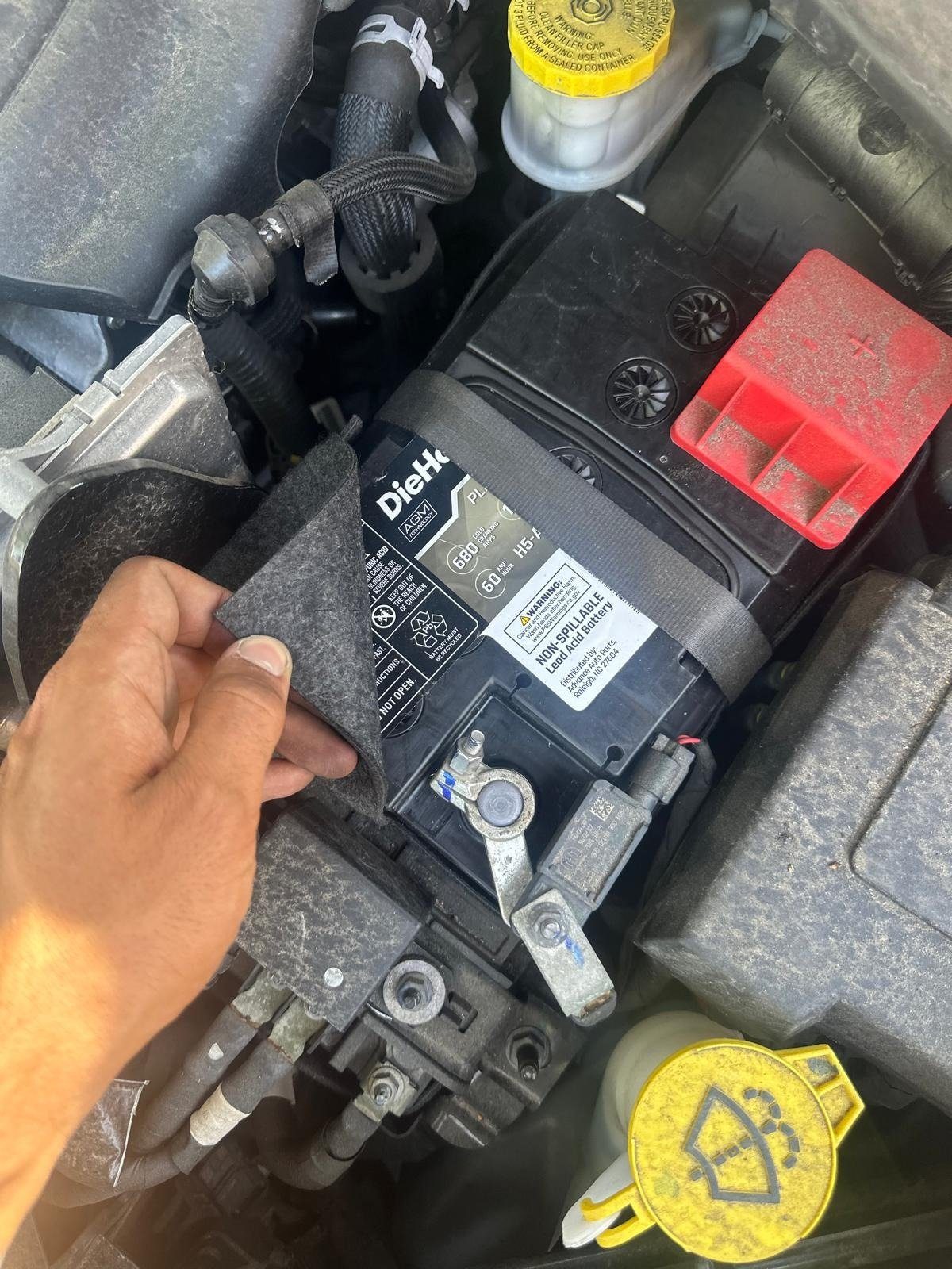 Technician lifting a felt cover on a DieHard AGM battery, showing the negative terminal clamp with a sensor and the red positive cover