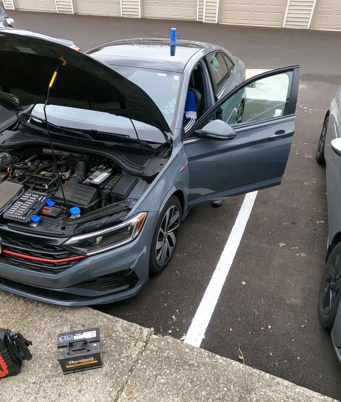 Volkswagen Jetta with its hood open in a parking lot during a mobile battery replacement, with the old battery on the curb
