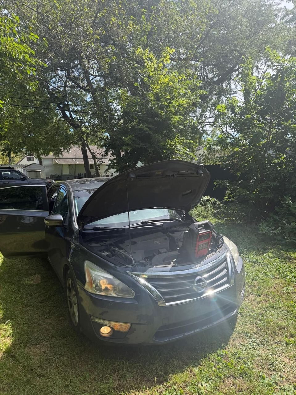 Nissan Altima with the hood open and a portable jump pack on the engine, parked on grass