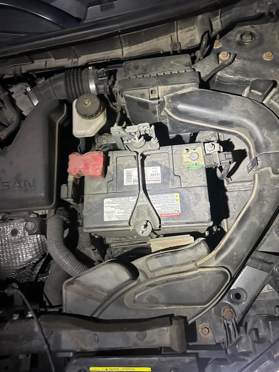 Car battery held down by a bracket across its top in a Nissan engine bay, red cover on the positive terminal