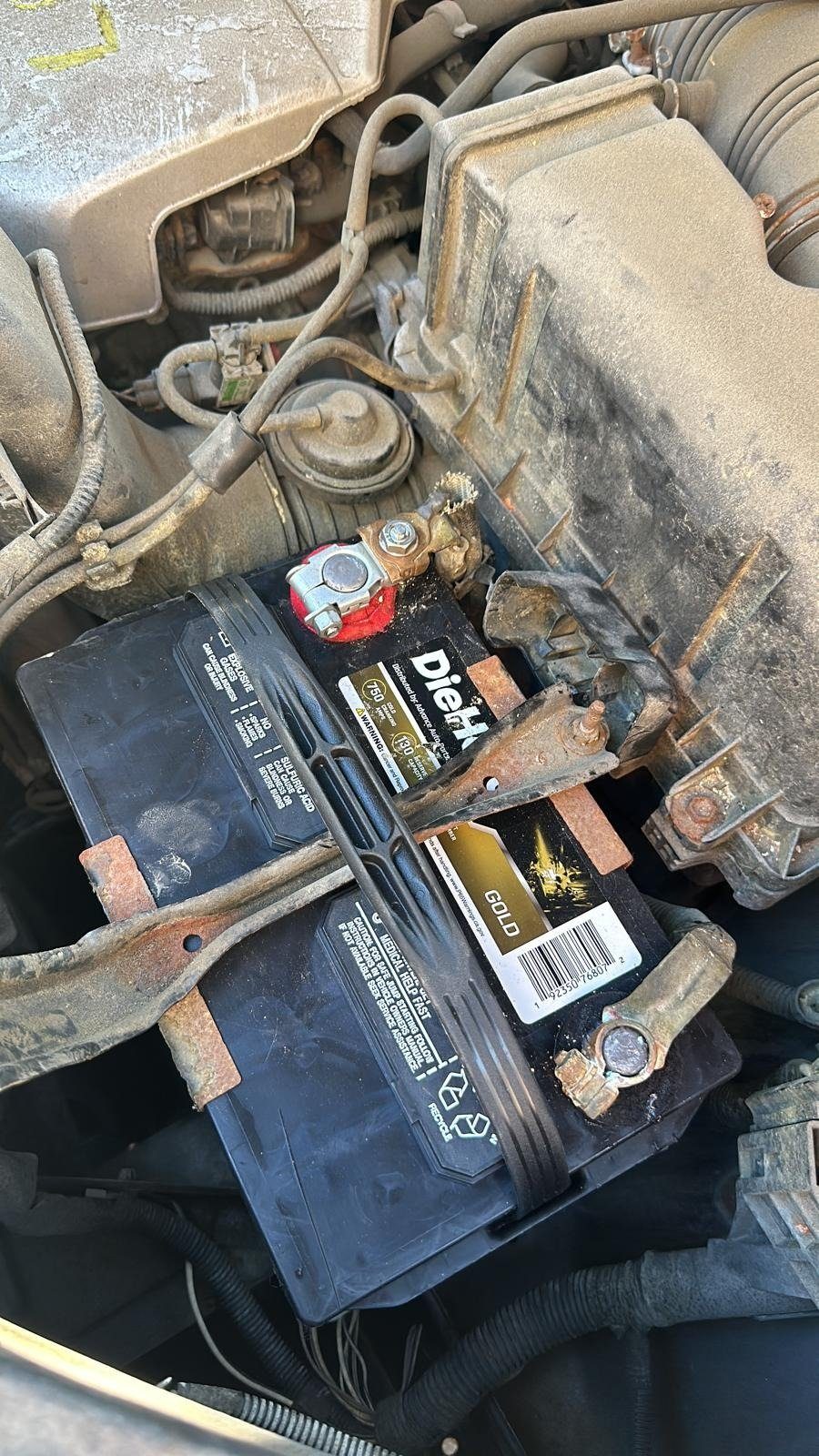 Old car battery in an engine bay with corroded terminal clamps and a rusted hold-down bracket