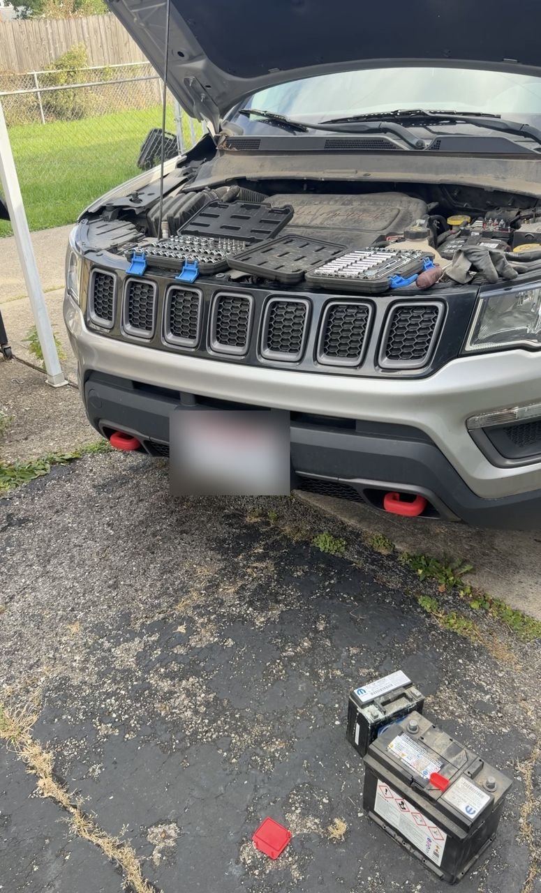 Gray Jeep Compass with the hood open and tool trays on the engine, the old main battery and the small auxiliary battery on the driveway