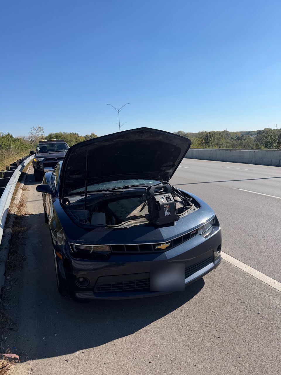 Dark blue Chevrolet Camaro with the hood up on a highway shoulder and a portable jump pack sitting on the engine, a service truck parked behind
