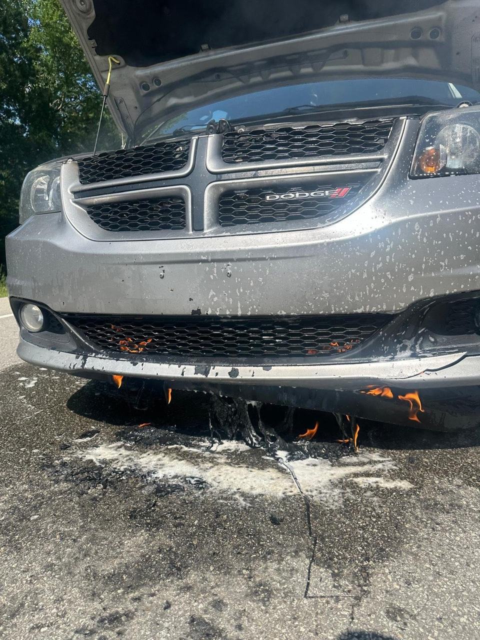 Small flames under the front bumper of a silver minivan, with extinguisher residue on the pavement