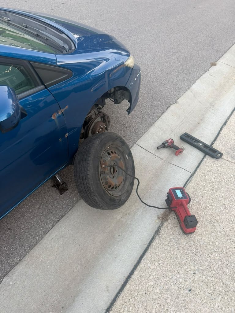 Blue sedan on a jack at the curb with its wheel off and a steel spare being inflated with a portable inflator