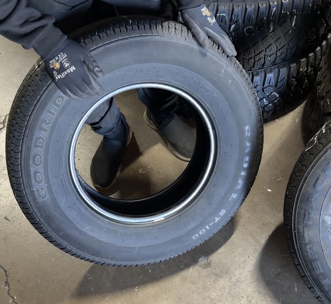 Gloved hands holding a used trailer tire marked ST225/75R15 at a tire shop, with more used tires stacked behind