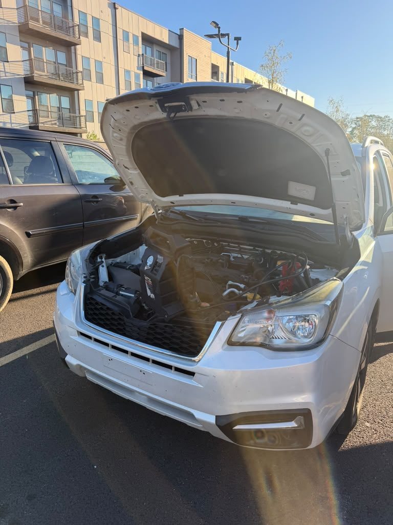 Portable jump pack clamped to the battery of a white Subaru Forester with its hood open in a parking lot