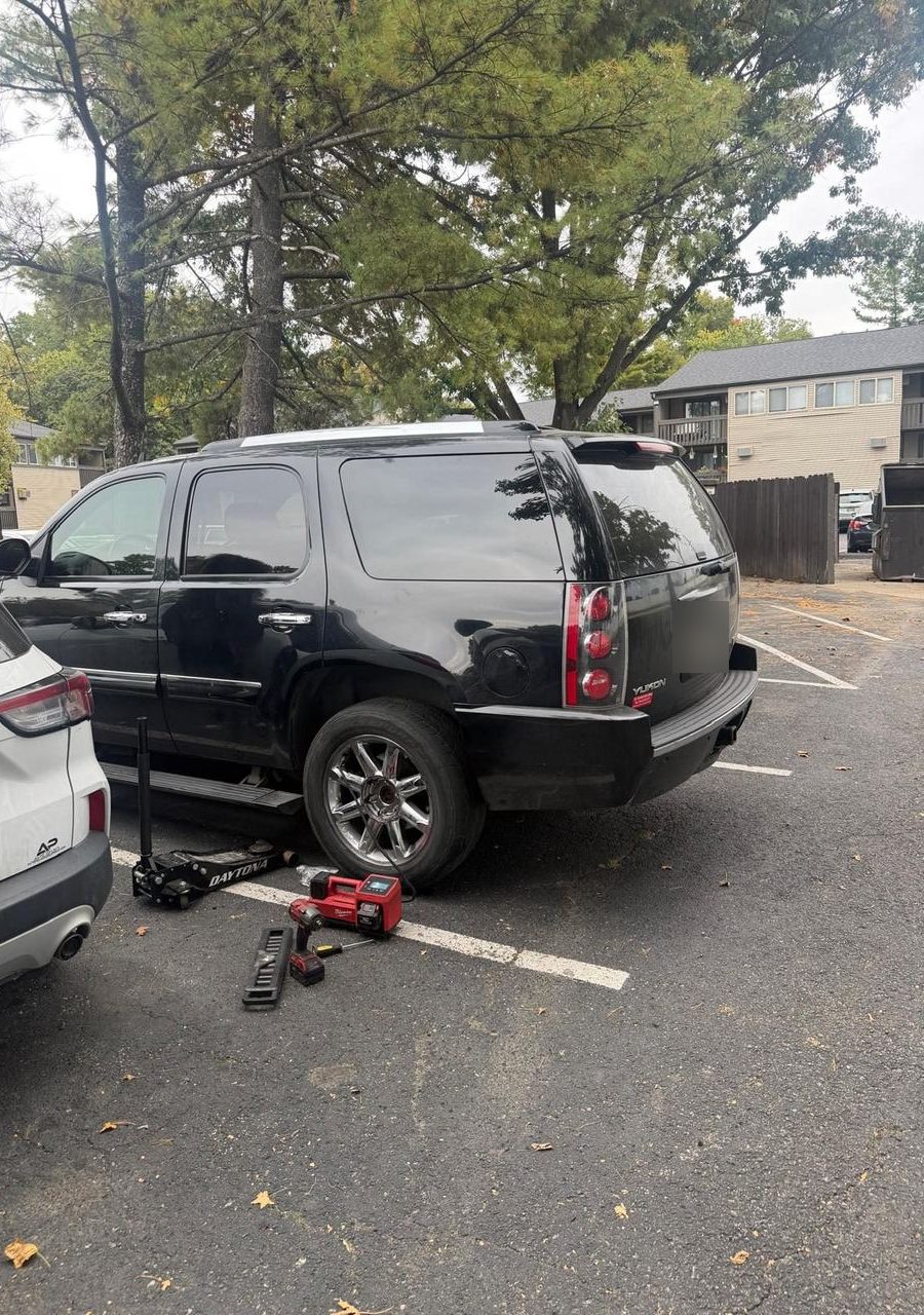 Black GMC Yukon in an apartment parking lot with a floor jack, impact wrench and portable inflator beside the rear wheel