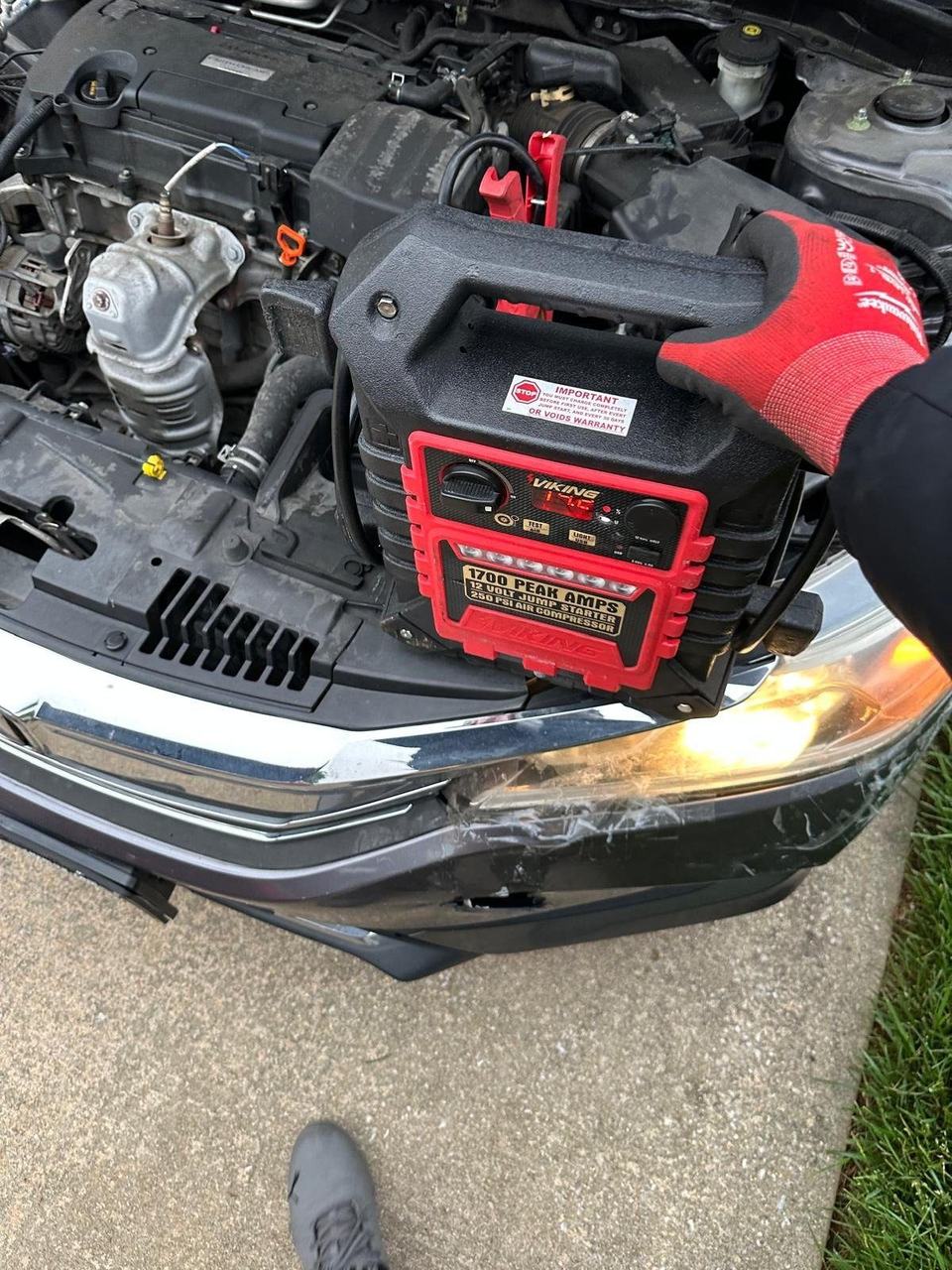 Technician in work gloves holding a 1700 peak amp jump starter in front of a Honda with the hood open
