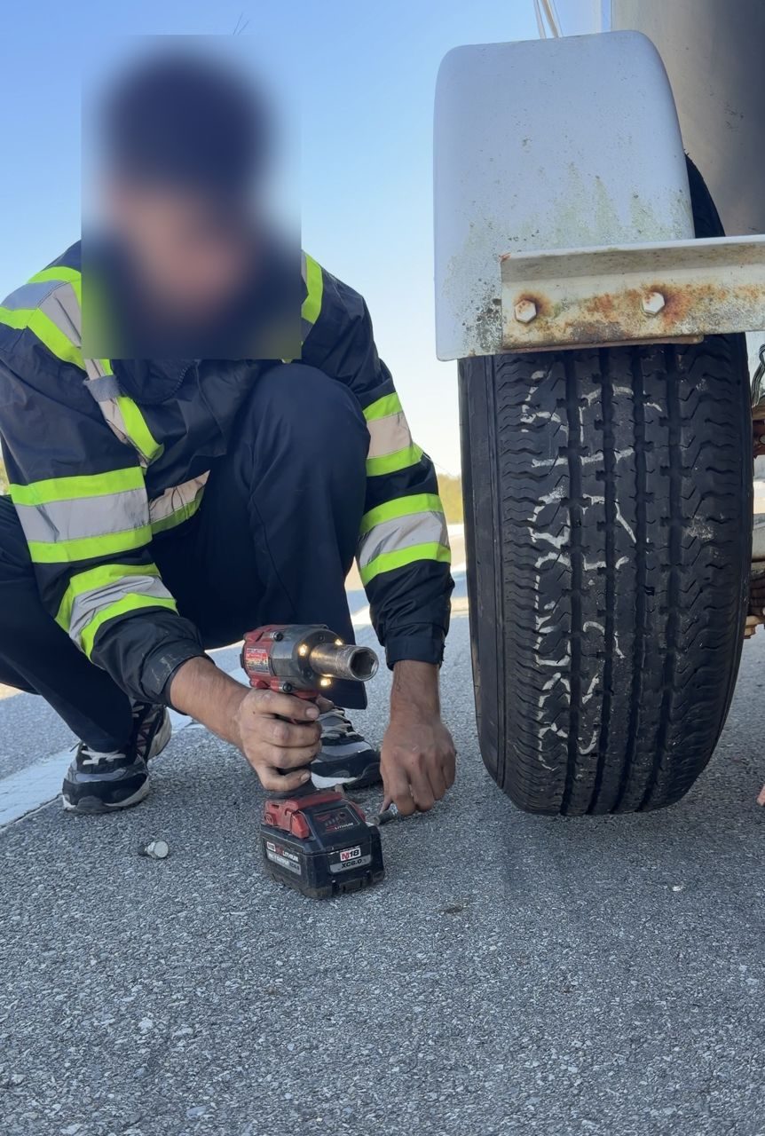 Technician in a high-vis jacket crouched by the boat trailer wheel with a cordless impact wrench on the highway shoulder
