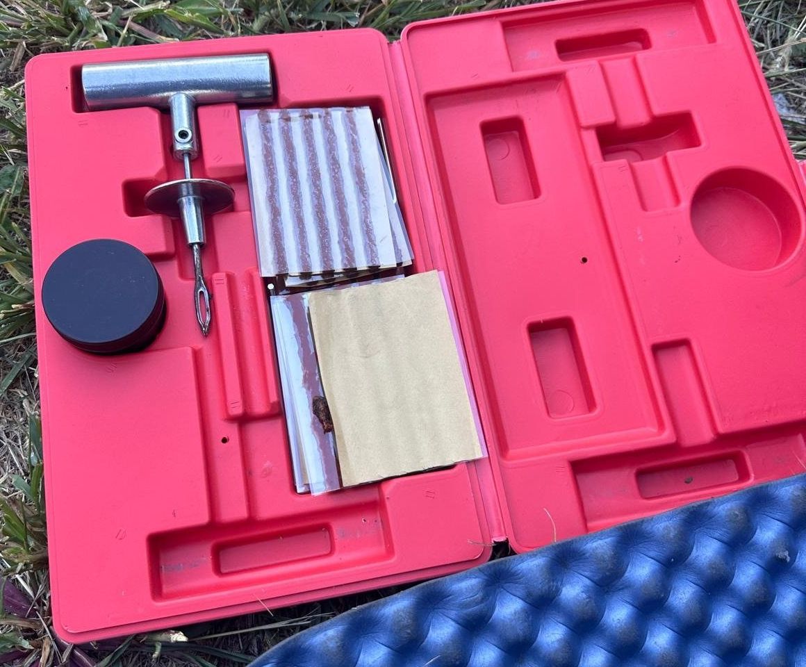 Open red tire repair kit on grass with a T-handle insertion tool, a strip of brown plug strings and a round tin