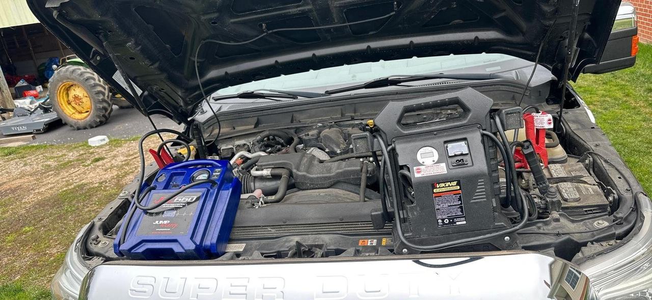 Two jump packs connected under the open hood of a Ford Super Duty pickup parked on grass