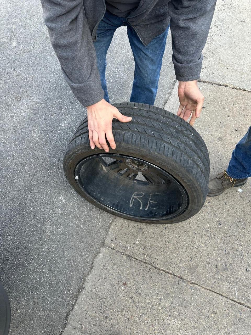 Person holding a wheel and tire taken off a car, the rim marked RF in chalk, on a sidewalk