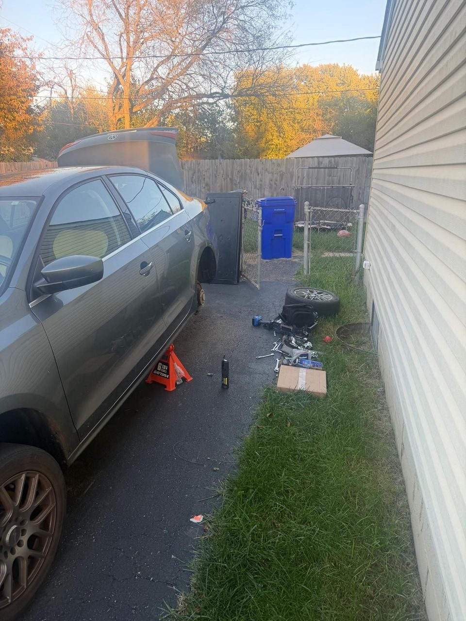 Grey sedan on a jack stand in a driveway with one wheel off and the tire and tools on the ground beside it